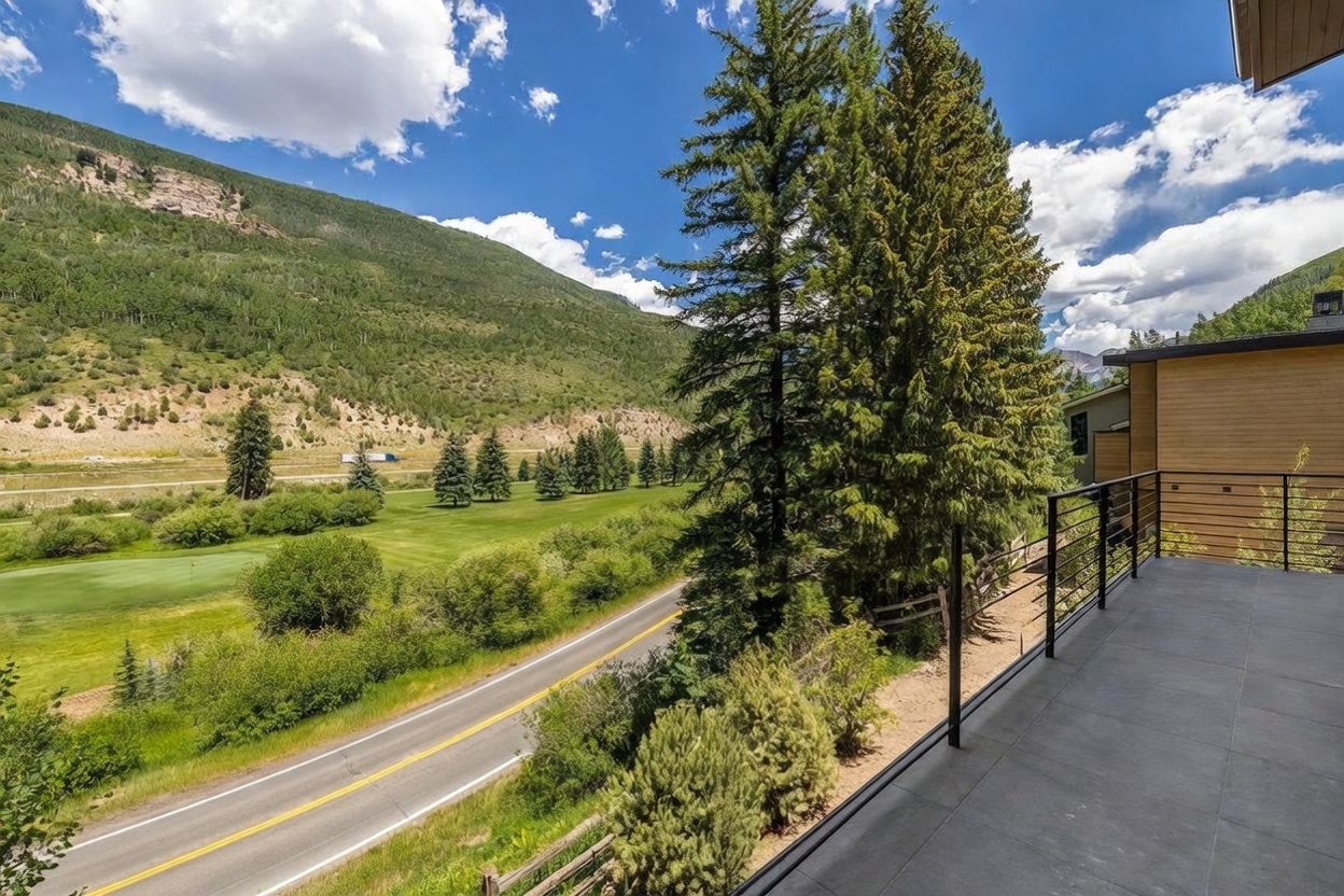This elevated balcony features modern metal railings and large-format grey tile flooring, offering a panoramic view of a lush green golf course and a winding road. The scene is framed by tall evergreen trees and a dramatic mountain range under a bright blue sky with scattered clouds. The perspective captures the seamless transition between the private outdoor living space and the expansive natural landscape.