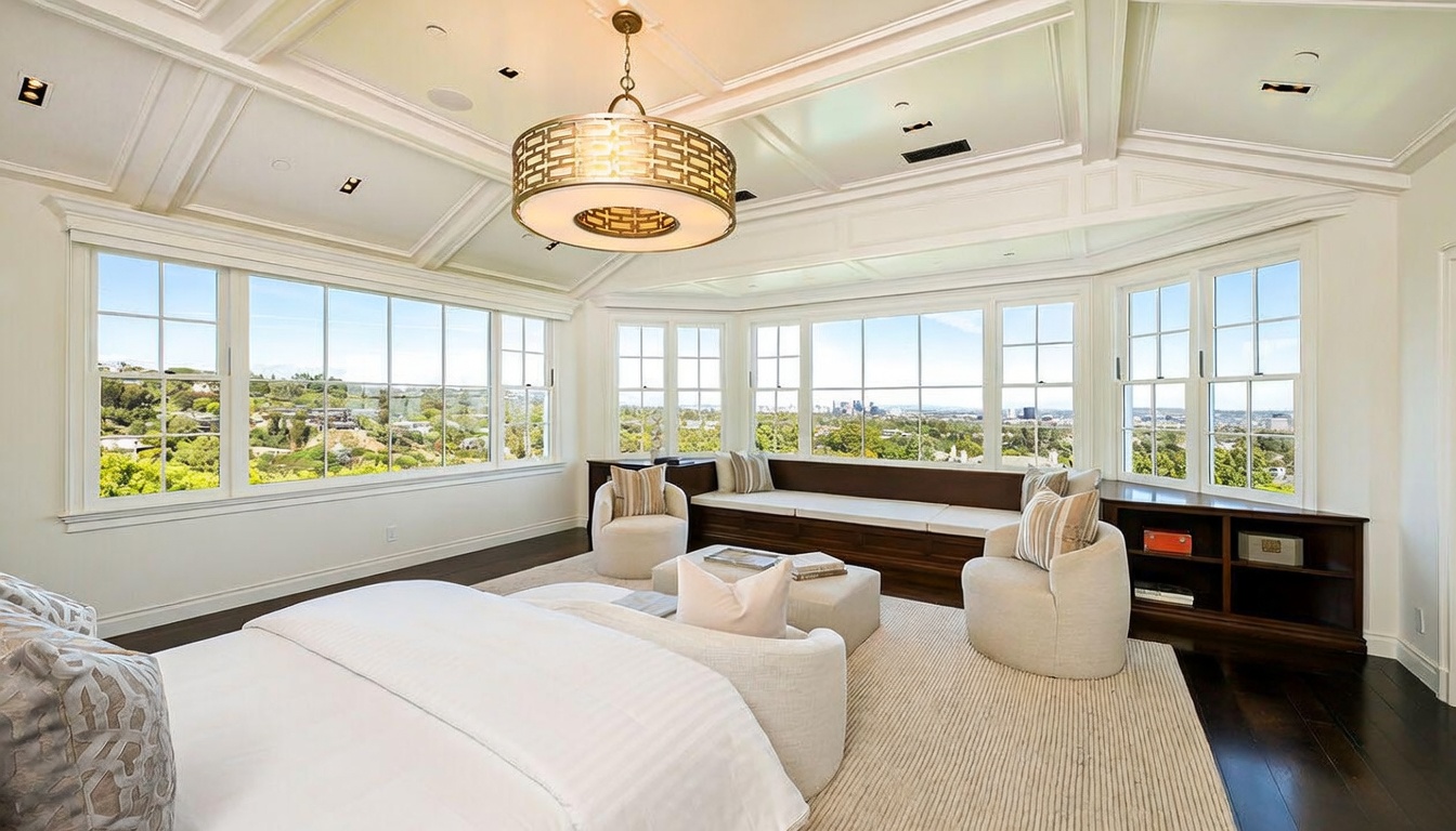 This spacious primary bedroom features a neutral color palette and a large bay window area that offers expansive views of the surrounding landscape and city skyline. The room is anchored by a plush bed and two comfortable armchairs, complemented by a built-in window seat with integrated storage. A decorative drum chandelier hangs from the coffered ceiling, adding a touch of elegance to the bright, airy space.