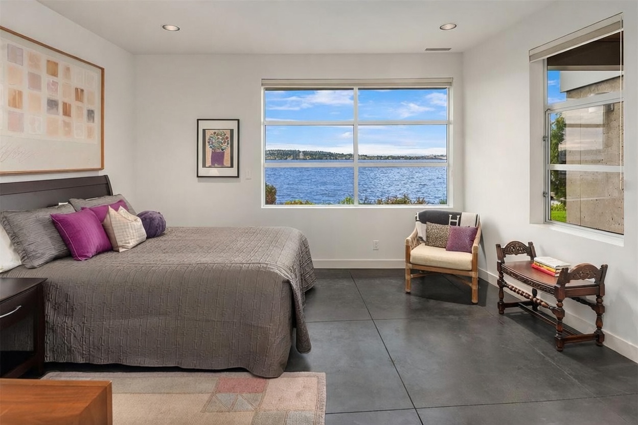 This primary bedroom features a modern aesthetic with large windows that frame a panoramic view of the lake. The room is furnished with a bed dressed in a grey quilted coverlet, accented by purple pillows, and includes a light-toned wooden chair and a decorative wooden bench. The space is finished with dark, large-format tile flooring, creating a sleek and contemporary atmosphere.