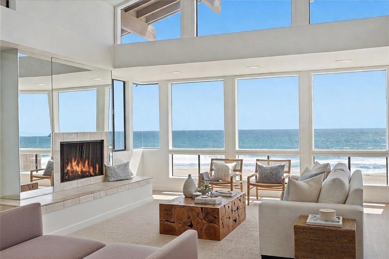 This bright and airy living room features a neutral color palette and a prominent fireplace with a stone-clad chimney breast. Large windows provide expansive, unobstructed views of the ocean, filling the space with natural light. The room is furnished with comfortable seating, including a plush sofa and armchairs, arranged around a unique, textured wood coffee table.