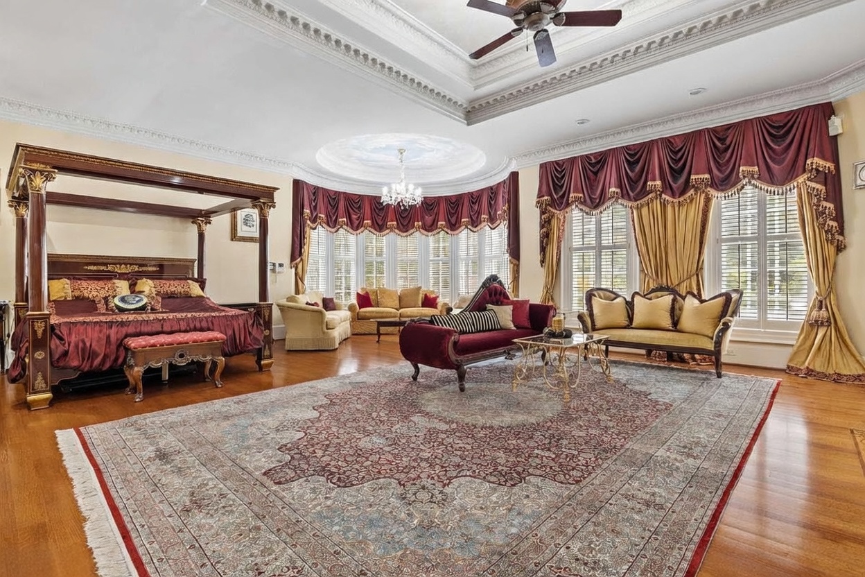 This expansive primary bedroom features a grand four-poster bed as the focal point, set against a backdrop of ornate, heavy drapery and a large bay window area. The room is decorated in a traditional, opulent style with a large, intricate area rug covering the hardwood floors and a central chandelier hanging from a tray ceiling. Multiple seating areas, including a velvet chaise lounge and a sofa, provide a luxurious and spacious feel to the suite.