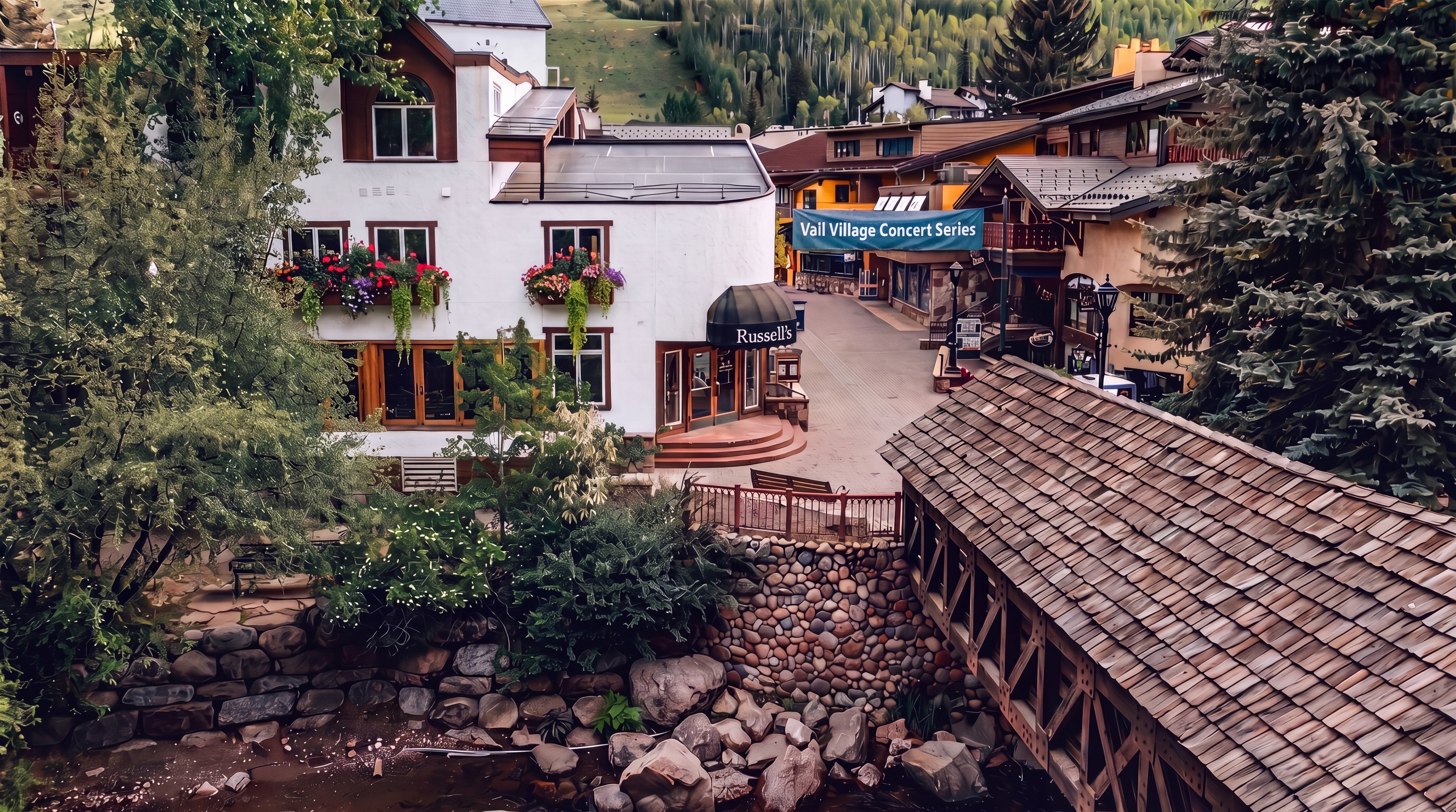 This elevated perspective captures a charming mountain village scene, featuring a rustic wooden covered bridge spanning a rocky stream in the foreground. The mid-ground showcases a multi-story building with a light-colored facade and dark trim, situated near a pedestrian walkway lined with shops and trees. The background reveals a lush, green hillside, creating a picturesque and inviting atmosphere typical of a resort destination.