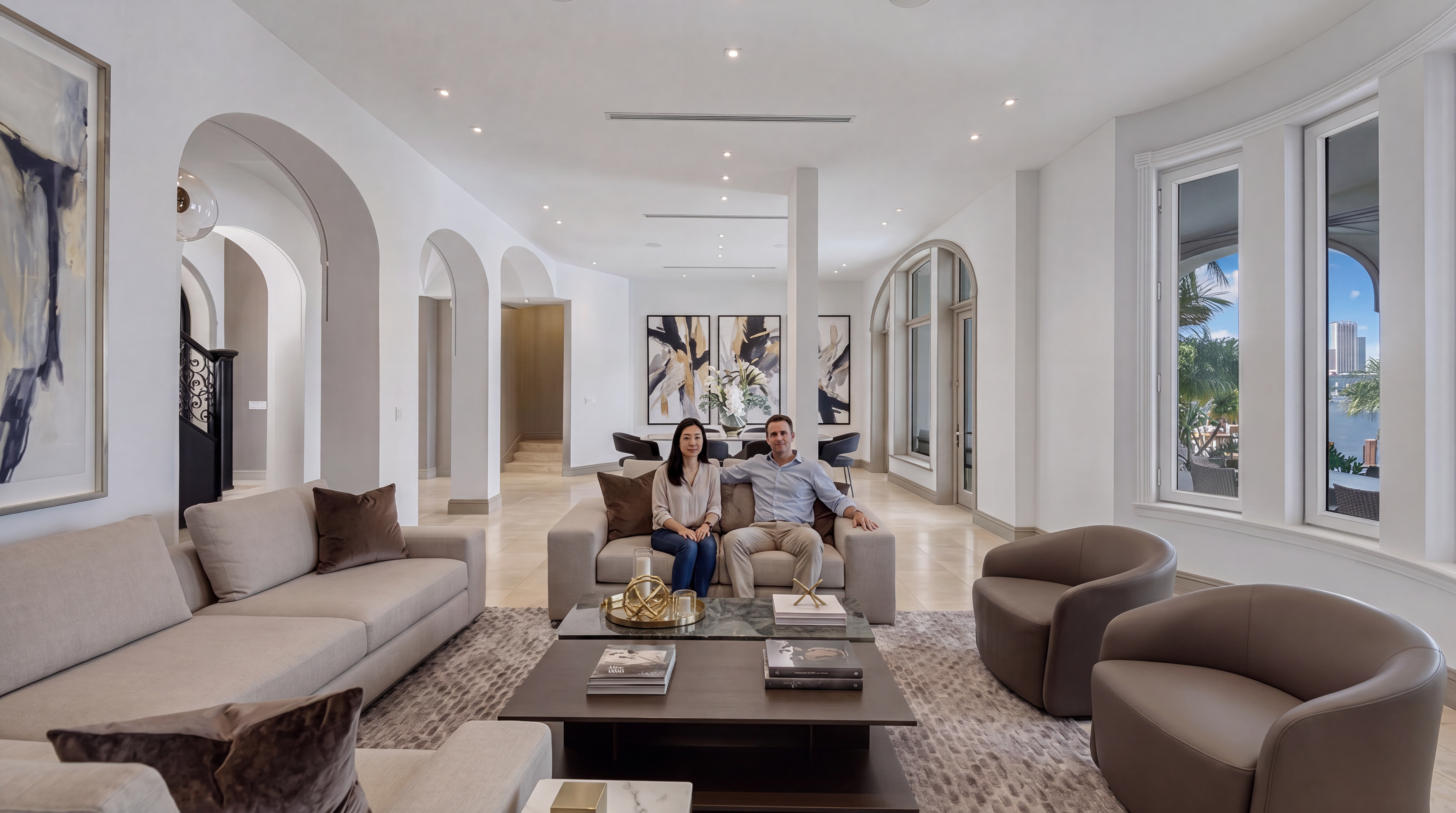 This spacious and elegant living room features a sophisticated neutral color palette, characterized by light beige sofas, comfortable lounge chairs, and a sleek, multi-tiered coffee table. The open-concept design is highlighted by graceful architectural archways, polished light-colored flooring, and large windows that offer scenic water views. The overall impression is one of refined luxury and harmonious, contemporary design.