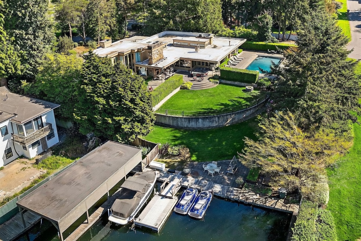 This high-angle aerial view captures a sprawling, modern waterfront estate nestled among mature trees. The property features a flat-roofed residence with a stone-clad chimney, a manicured lawn with a curved retaining wall, a swimming pool, and a private dock equipped with a boat slip and two jet skis. The lush landscaping and direct water access highlight the luxurious and private nature of this lakeside retreat.