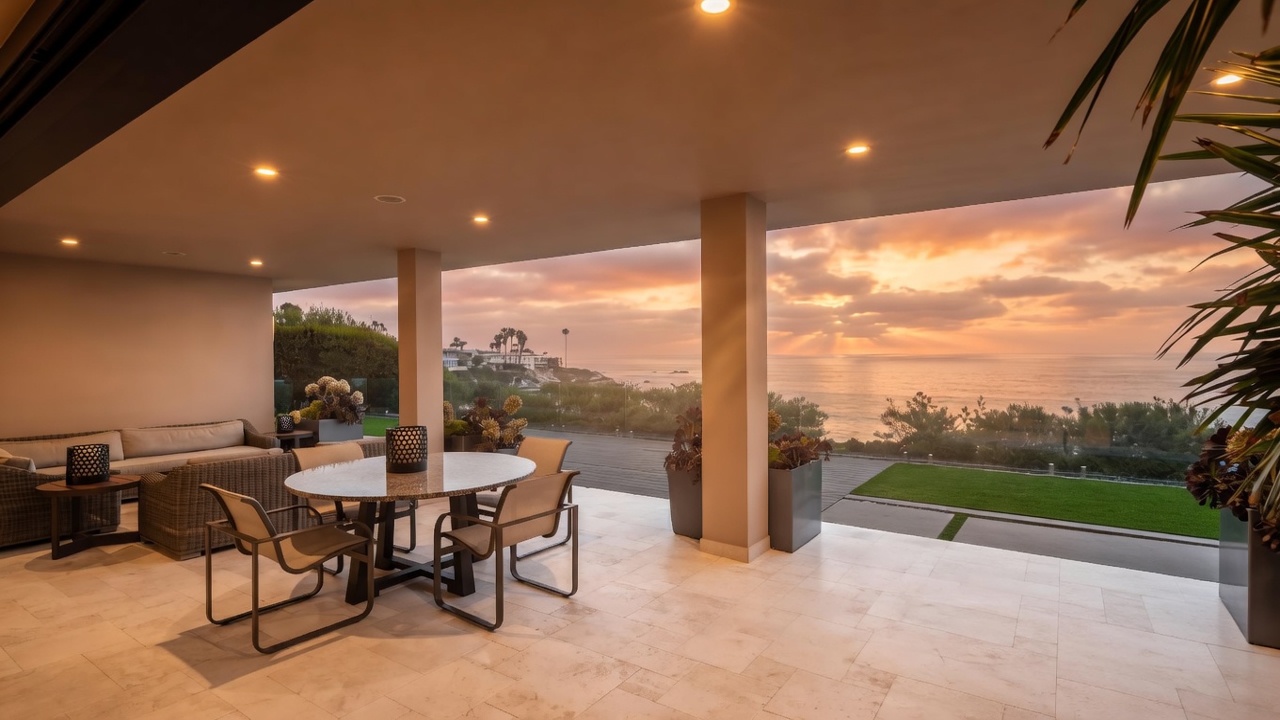 This expansive covered patio offers a seamless transition to an outdoor living space with a stunning, unobstructed ocean view. The area is furnished with a contemporary dining set and comfortable lounge seating, all set against a backdrop of a manicured lawn and the horizon. The open-air design and neutral stone flooring create a sophisticated, tranquil atmosphere perfect for coastal entertaining.