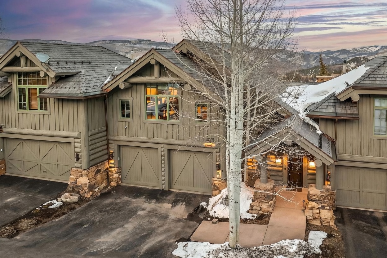 This elevated perspective captures the front facade of a multi-unit mountain residence, showcasing a rustic architectural style with dark-toned vertical siding and stone masonry accents. The structure features multiple gabled rooflines, a central garage area, and a welcoming entryway framed by stone pillars, all set against a backdrop of snow-dusted mountains at dusk. A prominent, leafless aspen tree stands in the foreground, adding a natural element to the scene while the warm glow from the interior windows creates an inviting atmosphere.