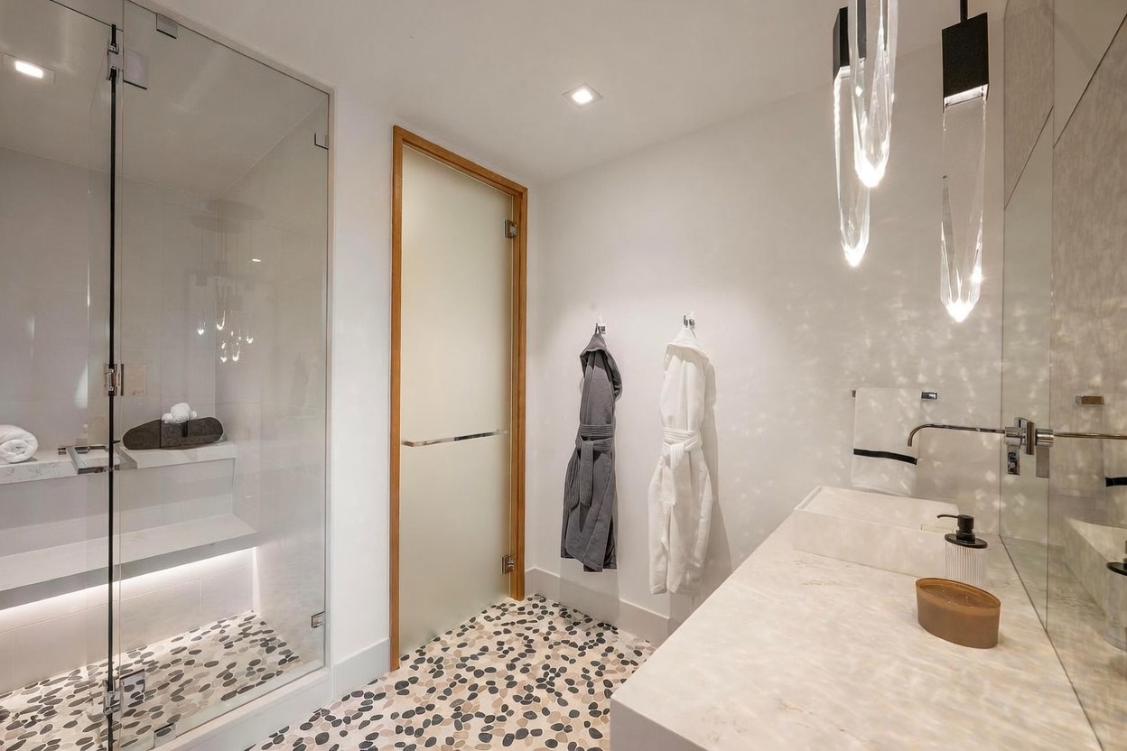 This modern bathroom features a walk-in shower with a glass enclosure and a built-in stone bench, complemented by a vanity with a stone countertop and a wall-mounted faucet. The floor is finished with a distinctive pebble-style tile, and two plush robes hang on the wall near a frosted glass door. The space is illuminated by sleek, sculptural pendant lights, creating a clean and sophisticated atmosphere.