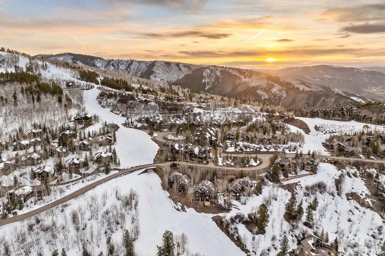 This expansive aerial view captures a luxury mountain community nestled within a snow-covered landscape during a vibrant sunset. The scene features numerous multi-story mountain-style residences with dark wood and stone exteriors, integrated seamlessly into the rugged, forested terrain. A winding road snakes through the development, which is positioned directly adjacent to groomed ski slopes, highlighting the prime ski-in/ski-out location.