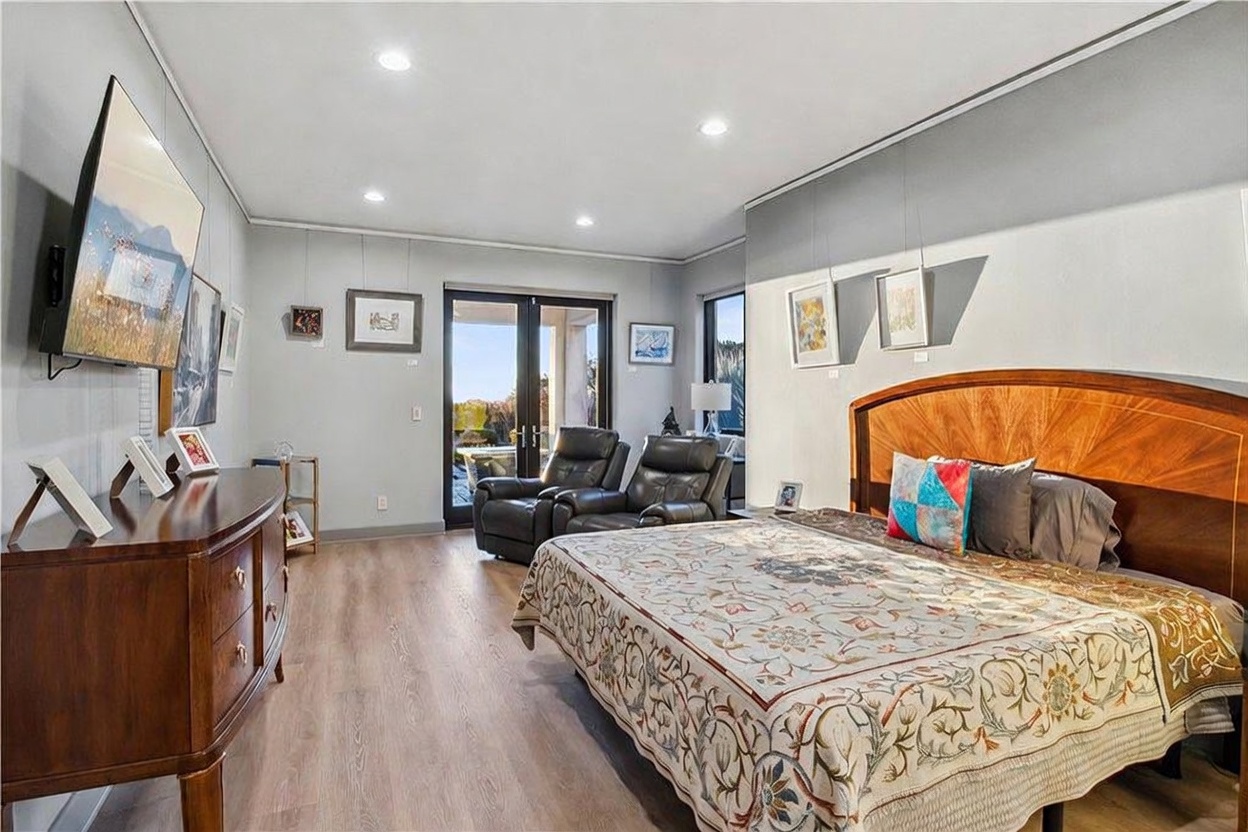 This spacious primary bedroom features light wood-look flooring and a large bed with a prominent curved wooden headboard. A dark wood dresser sits to the left, topped with a television, while two leather recliners are positioned near the glass doors that lead to an outdoor space. The room is decorated with various framed artworks on the walls and recessed lighting, creating a comfortable and well-lit living environment.