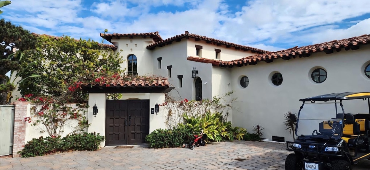 This Spanish-style home features a prominent gated courtyard entrance with a dark, paneled wooden door and a decorative tiled roof. The exterior is finished in a smooth, light-colored stucco, accented by circular windows and a golf cart parked on the stone-paved driveway. The scene is captured from a ground-level perspective, highlighting the architectural charm and private, gated access of the property.