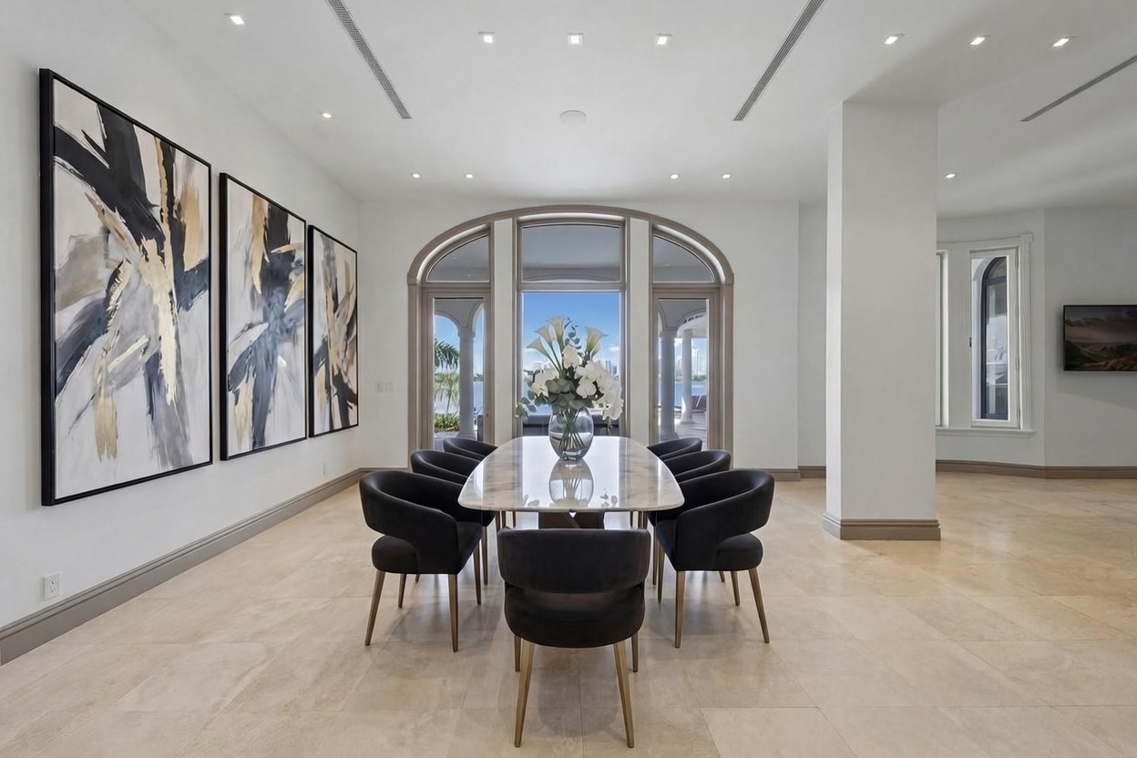 This elegant dining room features a long stone-topped table surrounded by eight dark upholstered chairs with metallic legs. The space is anchored by three large, abstract framed artworks on the left wall and a set of arched glass doors at the far end that provide a view of the water. The room is finished with light-toned stone flooring and recessed ceiling lighting, creating a sophisticated and airy atmosphere.