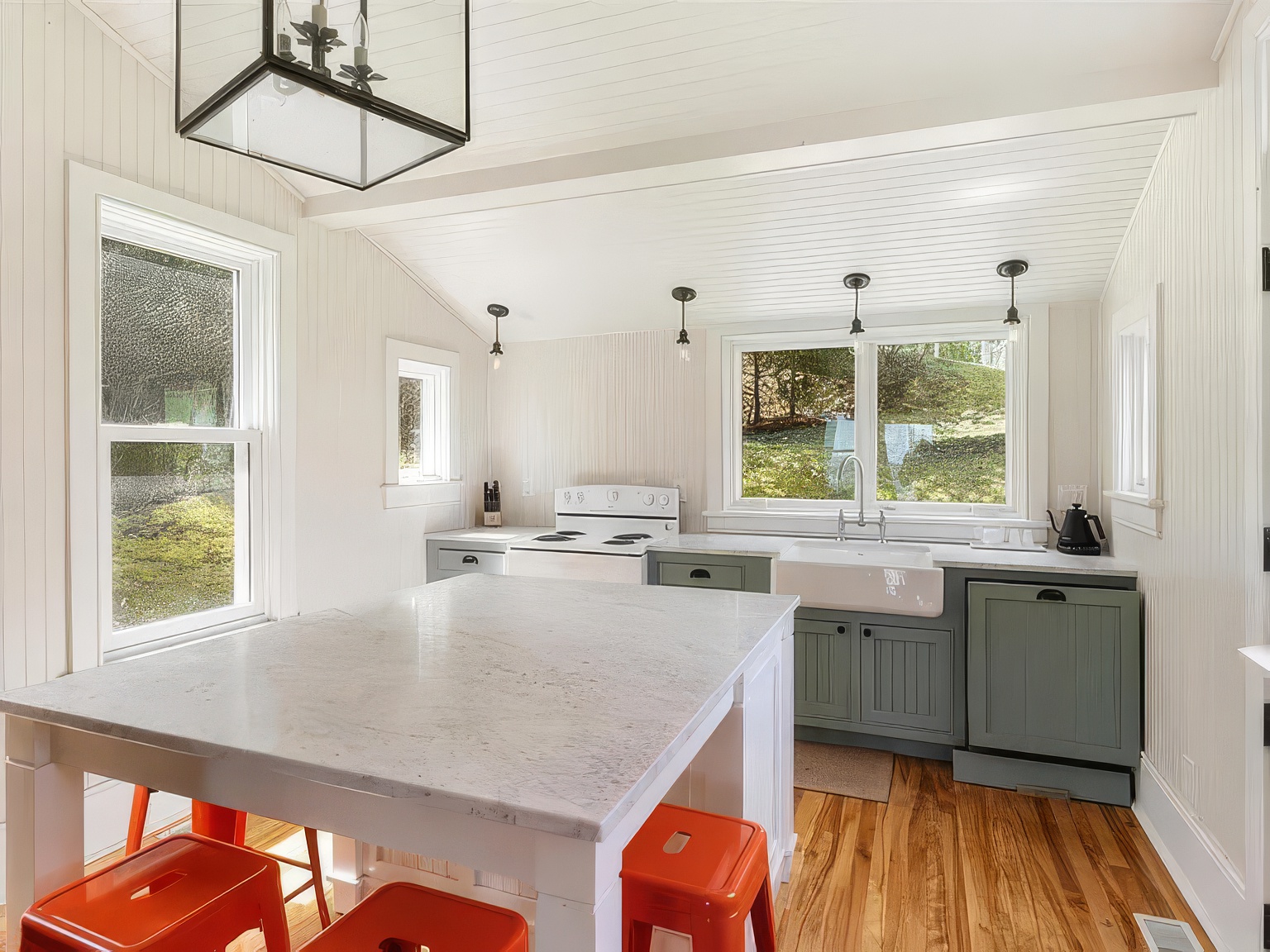 This bright, farmhouse-style kitchen features white beadboard walls, sage green lower cabinetry, and a large central island topped with a light-colored stone surface. The space is illuminated by a large black lantern pendant light and several industrial-style hanging bulbs, while a farmhouse sink sits beneath a window overlooking the outdoors. The warm wood flooring and vibrant orange stools add a welcoming, cozy contrast to the clean, neutral palette.