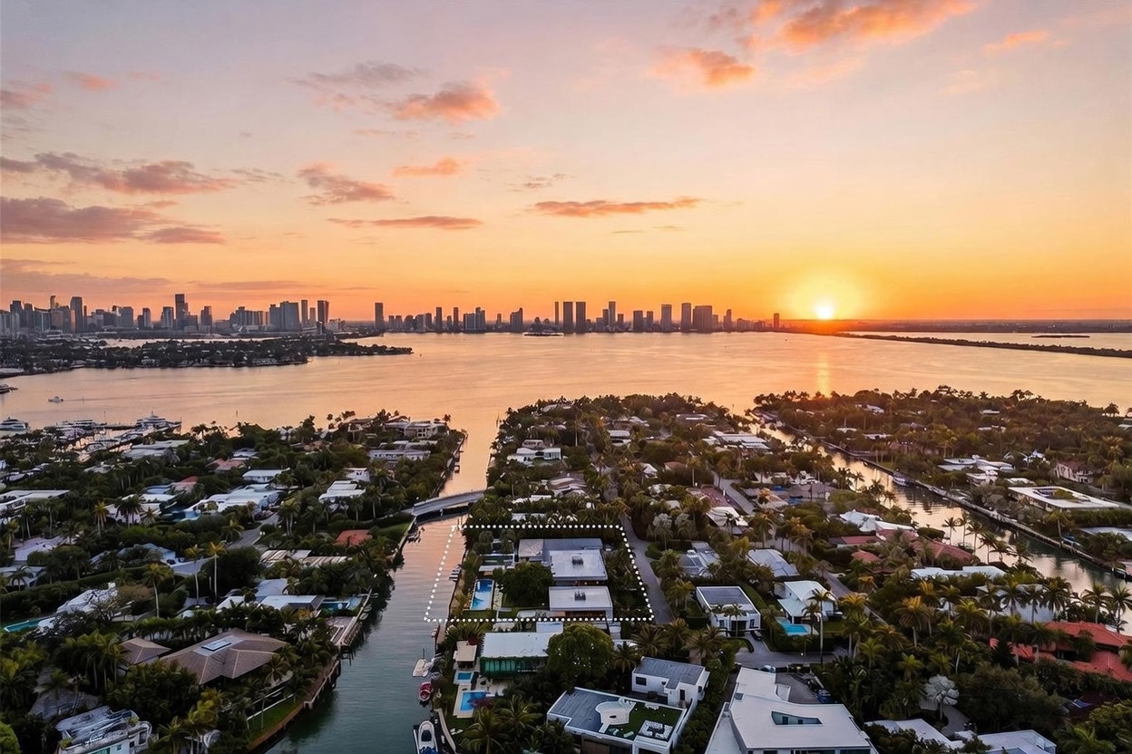 This high-angle aerial view captures a waterfront residential property at sunset, with a dotted outline highlighting the specific lot. The scene features a modern home with a swimming pool situated on a narrow peninsula, surrounded by lush tropical landscaping and calm canal waters. In the distance, a vibrant sunset illuminates the horizon behind a sprawling city skyline, creating a serene and luxurious atmosphere.