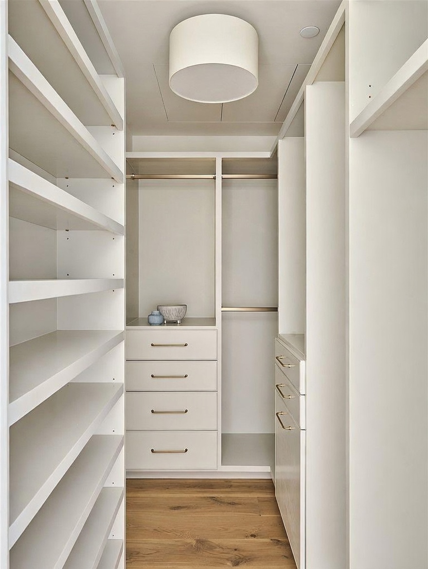 This walk-in closet features a clean, minimalist design with custom white cabinetry, including open shelving on the left and a combination of hanging rods and drawers on the right. The space is illuminated by a central drum-style ceiling light and features warm-toned wood flooring that adds a touch of natural texture. The symmetrical layout and neutral color palette create an organized and sophisticated storage environment.
