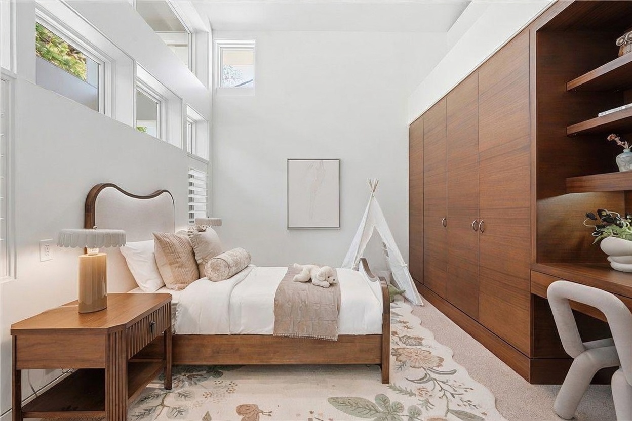 This bright and airy bedroom features high ceilings with a series of clerestory windows that flood the space with natural light. The room is furnished with a wooden bed frame, a matching nightstand with a decorative lamp, and a built-in wood-toned wardrobe unit that incorporates a desk area. A whimsical play tent and a floral-patterned area rug add a playful, cozy touch to the modern, minimalist design.