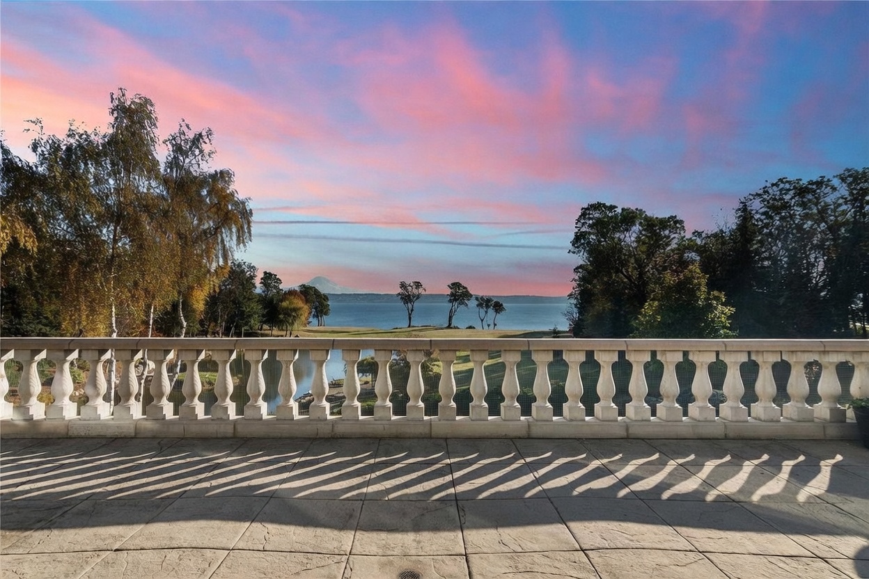 This elevated balcony features a classic stone balustrade that overlooks a sprawling lawn, a calm body of water, and a distant mountain peak under a vibrant, pink-hued sunset sky. The stone-tiled floor creates long, dramatic shadows, emphasizing the depth and peaceful atmosphere of this outdoor living space. The perspective is wide and cinematic, capturing the serene transition from the private terrace to the expansive natural landscape.