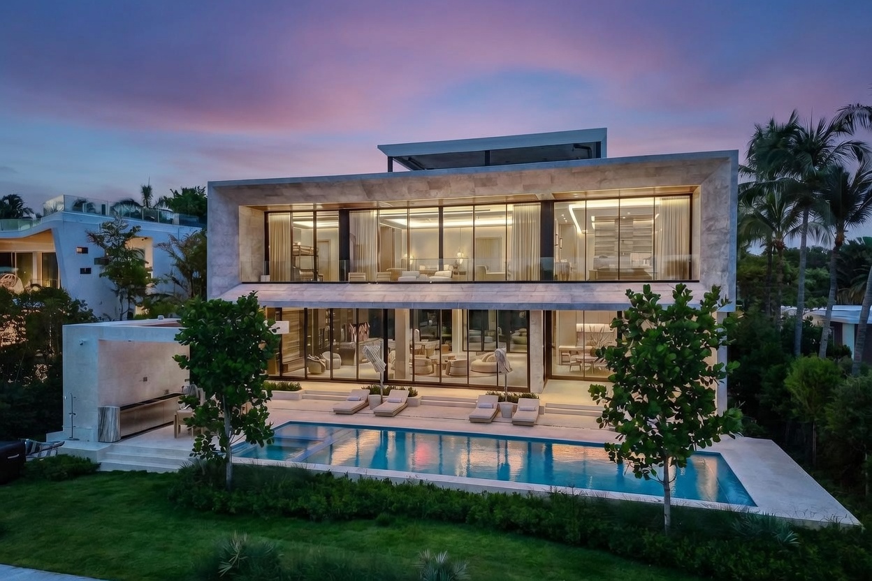 This stunning contemporary residence features a striking two-story design with expansive floor-to-ceiling glass walls that seamlessly connect the interior living spaces to the outdoors. The rear exterior showcases a sleek, rectangular swimming pool set within a manicured lawn, complemented by a stone-paved patio area furnished with lounge chairs. The architectural style is defined by clean lines, stone cladding, and a sophisticated, modern aesthetic captured during the twilight hour.