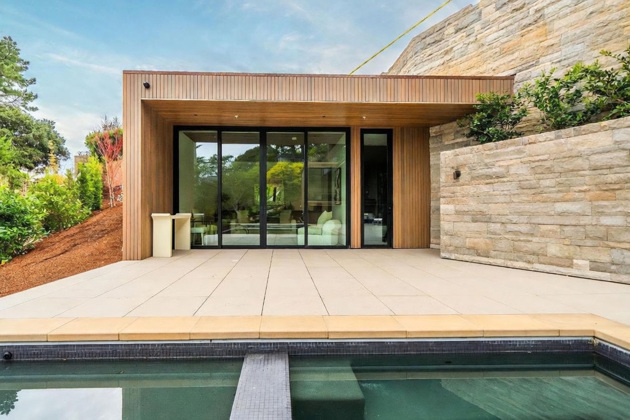 This modern outdoor space features a sleek, rectangular swimming pool in the foreground with a stone-tiled patio leading to a contemporary structure. The building showcases warm-toned wood paneling and large glass sliding doors that provide a seamless transition between the indoor and outdoor living areas. A textured stone wall adds architectural interest, while the surrounding landscape includes a sloped, mulched garden area.