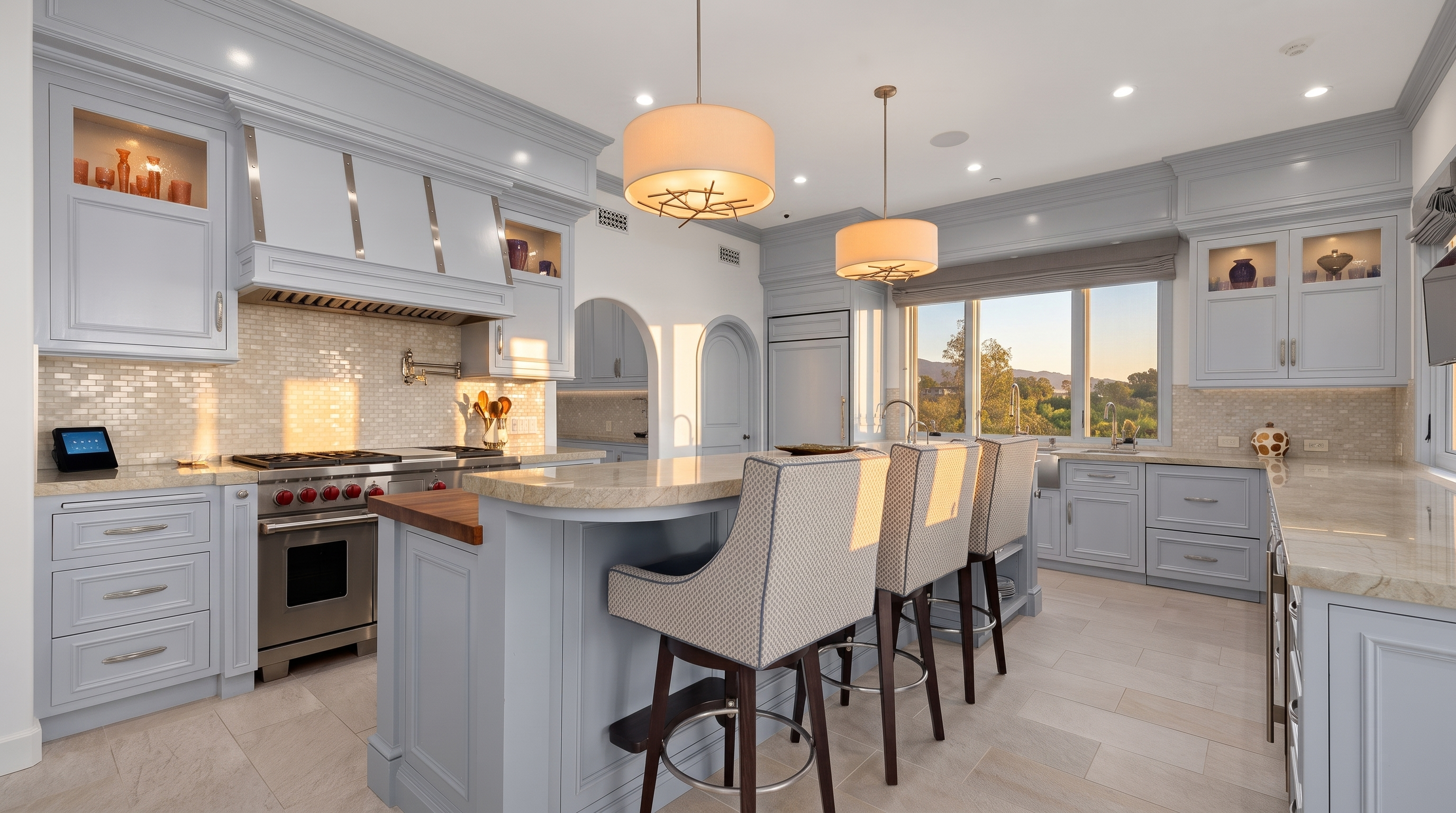 This bright and spacious kitchen features light blue cabinetry paired with neutral stone countertops and a matching tiled backsplash. A large central island with a wood-topped section provides seating for three, illuminated by two decorative pendant lights. The room is anchored by a professional-grade stainless steel range and offers a pleasant view of the outdoors through a wide window above the sink.