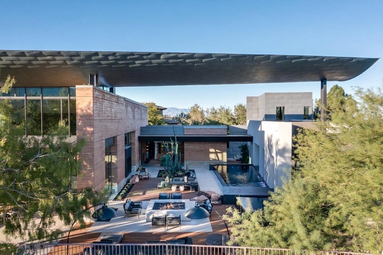 This high-angle aerial perspective captures a luxurious modern residence featuring a sprawling outdoor living space sheltered by a massive, cantilevered roof structure. The area includes a rectangular swimming pool, a central fire pit surrounded by lounge seating, and a desert-inspired landscape with mature cacti. The architecture emphasizes clean lines, stone-clad walls, and expansive glass, creating a seamless transition between the indoor and outdoor environments.