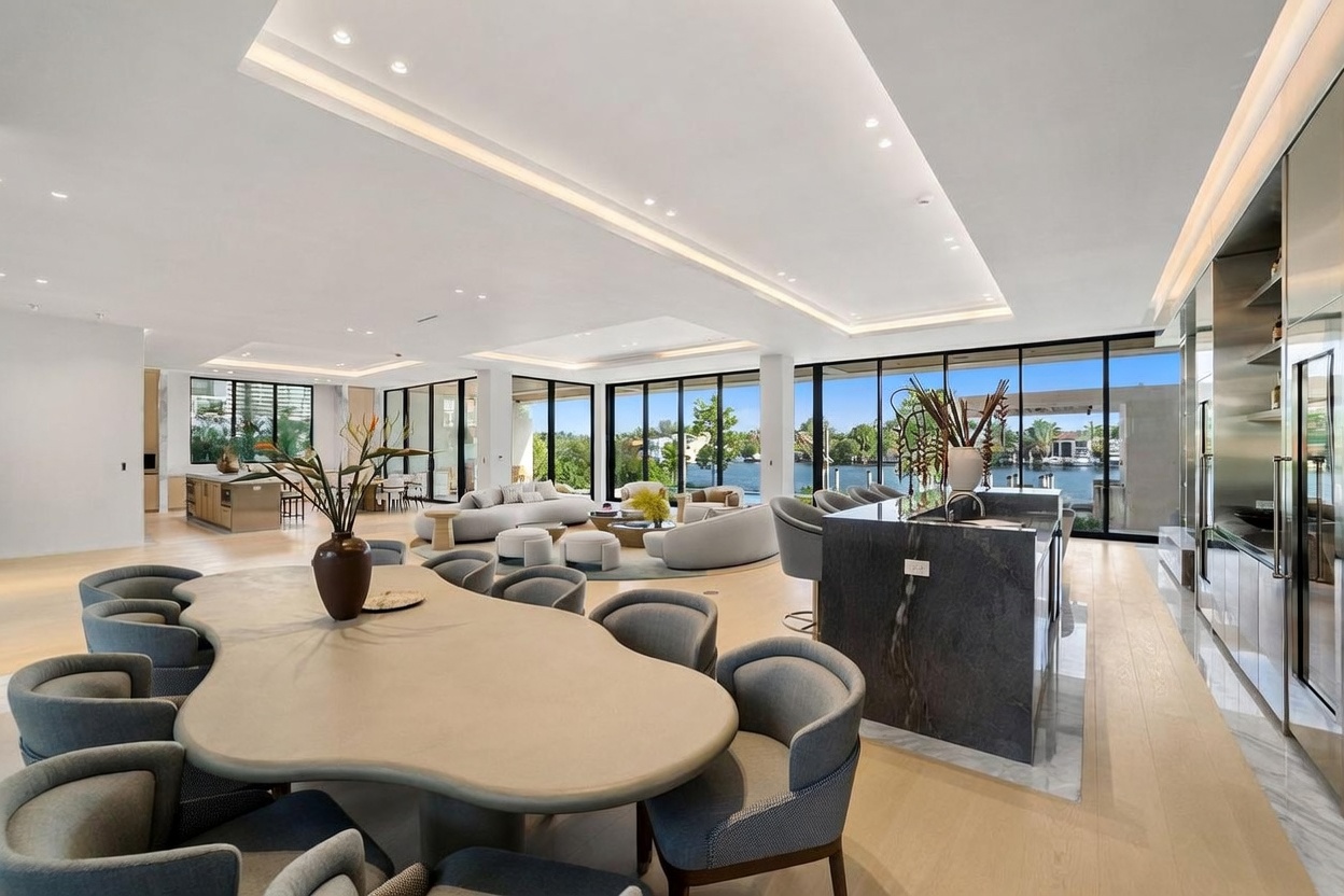 This expansive, open-concept living and dining area features a sophisticated, modern aesthetic with light wood flooring and a neutral color palette. The space is anchored by a large, uniquely shaped dining table in the foreground and a comfortable seating area in the background, all framed by expansive glass walls that offer a clear view of the waterfront. The room is illuminated by recessed lighting and sleek, minimalist fixtures, creating a bright and airy atmosphere perfect for luxury entertaining.