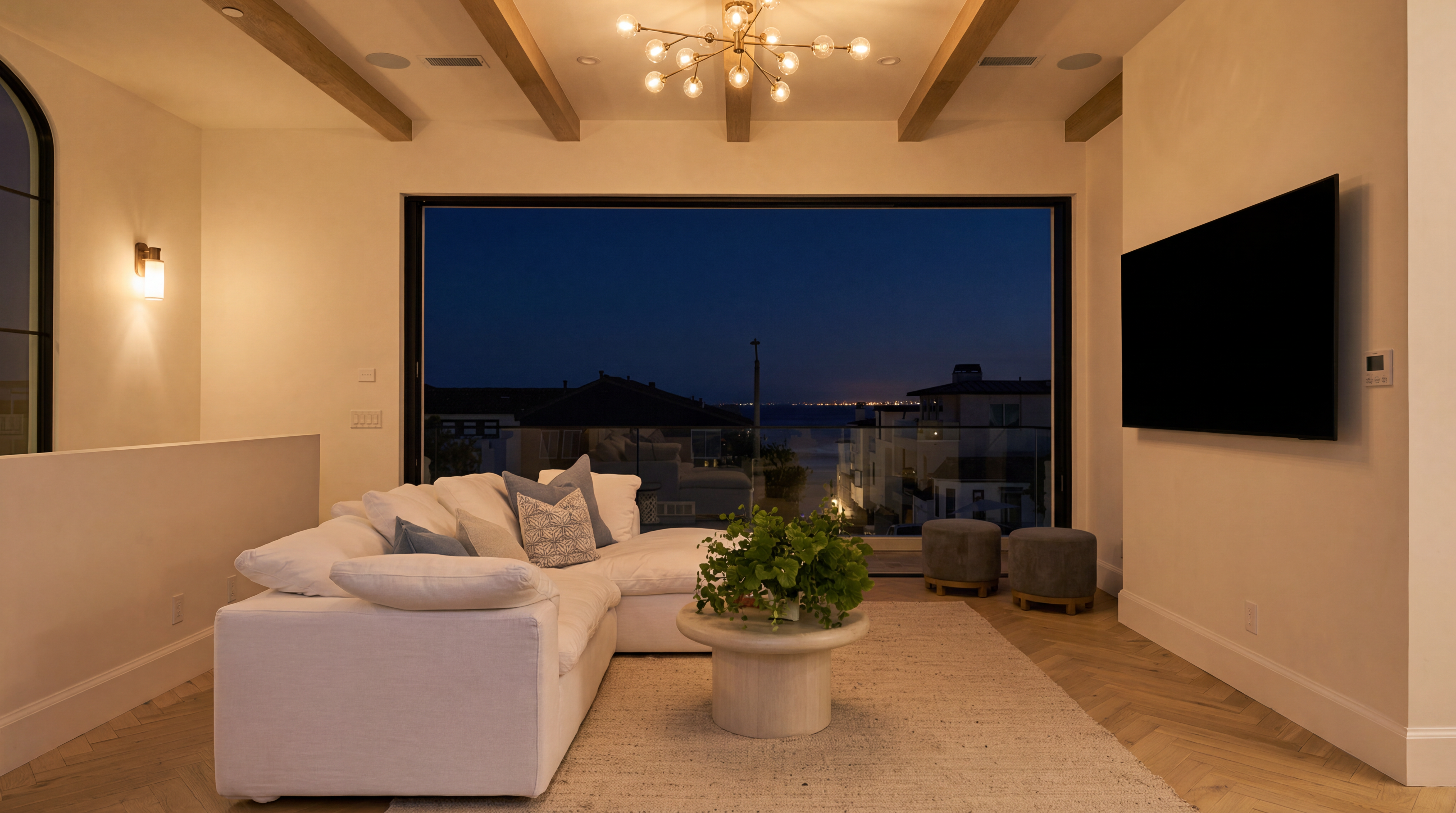 This sophisticated, contemporary living room features a spacious white sectional sofa, a textured area rug, and a modern drum-style coffee table adorned with a vibrant plant. The room is anchored by a large, picture-frame window that offers expansive evening views, complemented by warm wood ceiling beams and statement globe lighting. The minimalist design is further enhanced by a wall-mounted television, creating an inviting space perfect for both relaxation and entertaining.