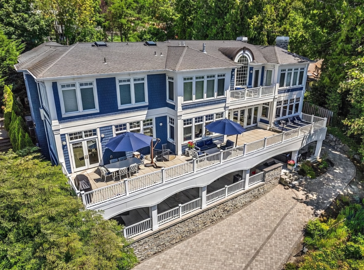 This high-angle exterior shot showcases the expansive rear facade of a multi-story, blue-shingled luxury home with white trim. The property features a massive, multi-level deck equipped with outdoor dining areas, lounge seating, and umbrellas, overlooking a manicured lawn and stone-paved walkway. The architectural design emphasizes indoor-outdoor living, with large windows and glass doors that provide a seamless transition to the elevated outdoor entertainment spaces.