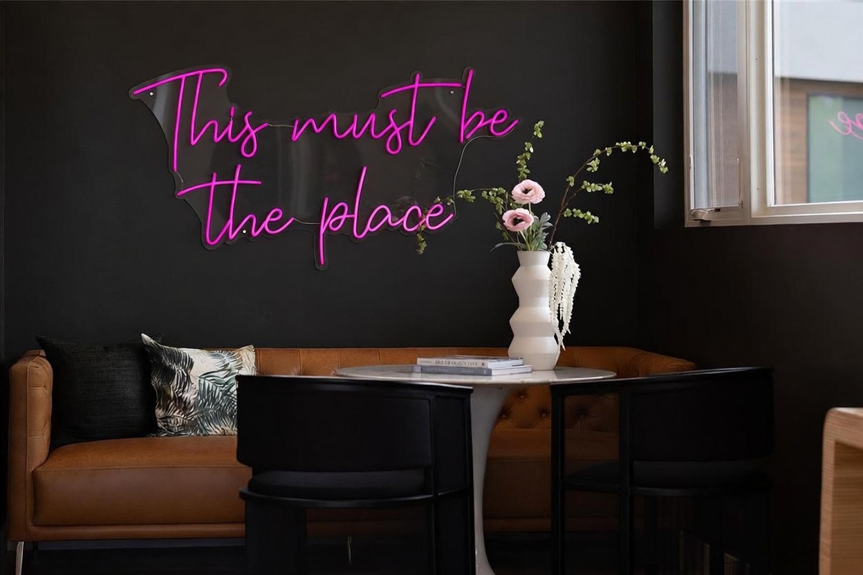This stylish dining nook features a tufted leather banquette set against a dramatic matte black wall adorned with a vibrant pink neon sign reading 'This must be the place.' A sleek, white pedestal table is centered in the space, accompanied by two modern black chairs and a decorative vase with floral accents. The composition offers a chic, intimate atmosphere perfect for casual dining or conversation.