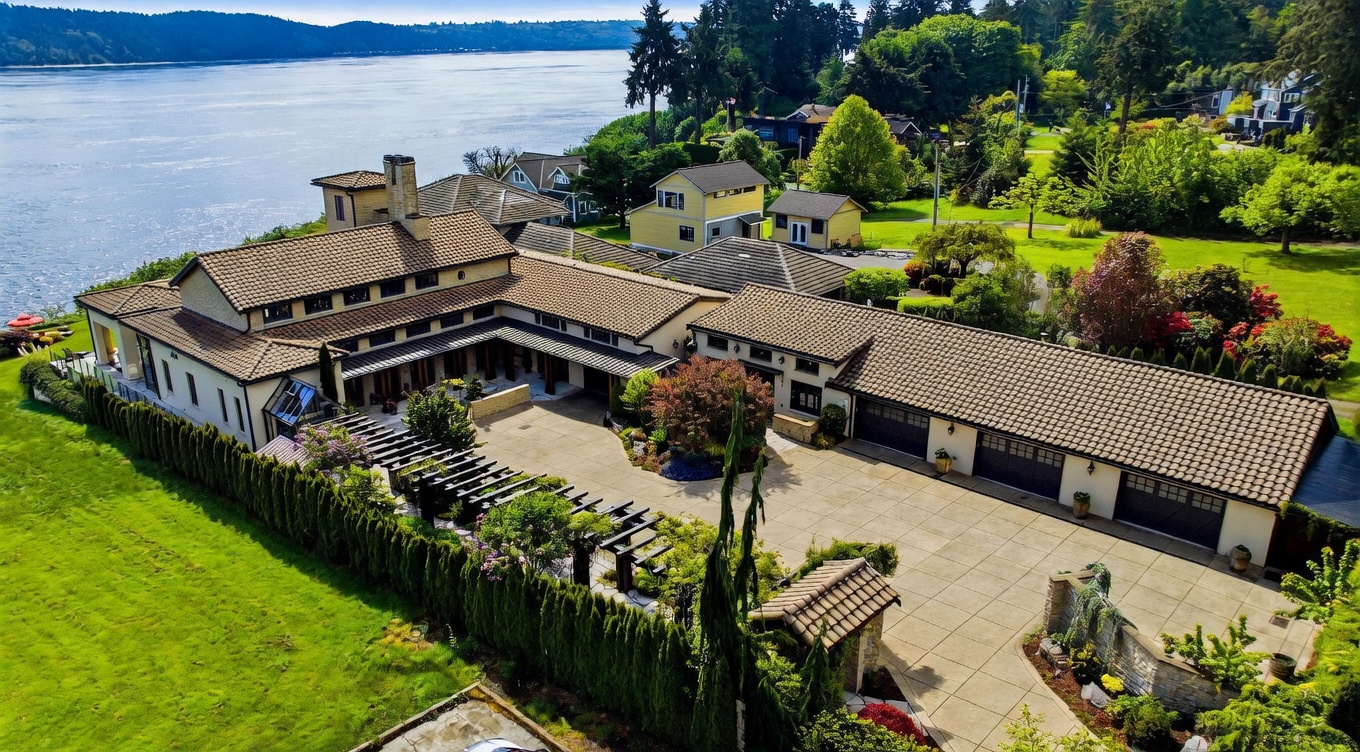 This high-angle aerial view showcases a sprawling, U-shaped estate situated directly along a scenic waterfront. The property features a large paved courtyard, a multi-car garage, and extensive manicured lawns, all framed by lush greenery and mature trees. The architecture is characterized by a classic Mediterranean-inspired style with tiled roofing and a cohesive, expansive layout that emphasizes indoor-outdoor living.