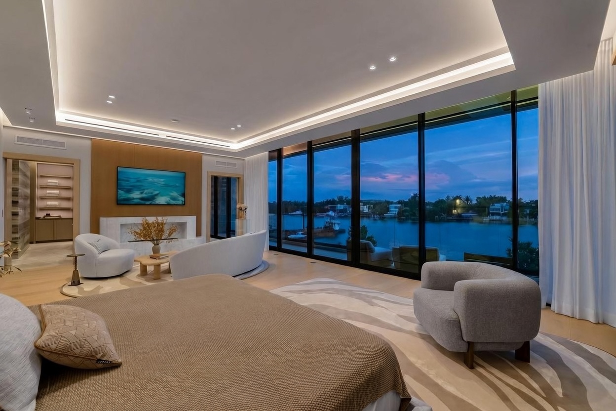 This expansive primary bedroom features a sophisticated, neutral color palette and a contemporary design. The room is anchored by a large bed in the foreground and a cozy seating area with a curved sofa and armchair, all oriented toward a wall-mounted television and a sleek fireplace surround. Expansive floor-to-ceiling windows provide a panoramic view of the waterfront at dusk, creating a serene and luxurious atmosphere.