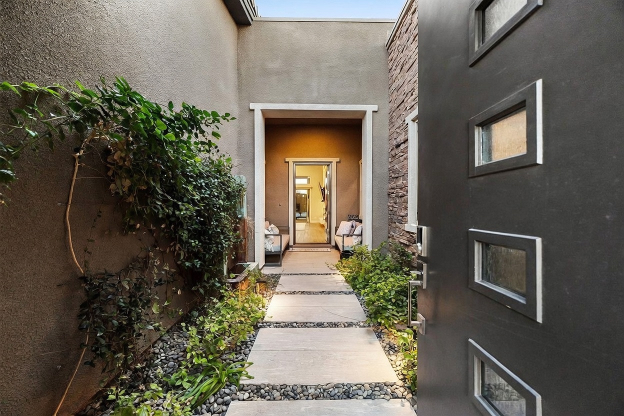 This perspective captures a modern, semi-enclosed entryway viewed from just inside a dark, paneled front door. A stone-paved walkway leads through a landscaped courtyard filled with river rocks and lush greenery toward a secondary glass door that reveals a bright interior space. The architectural design emphasizes a private, transitional outdoor foyer that creates a sophisticated and welcoming transition into the home.