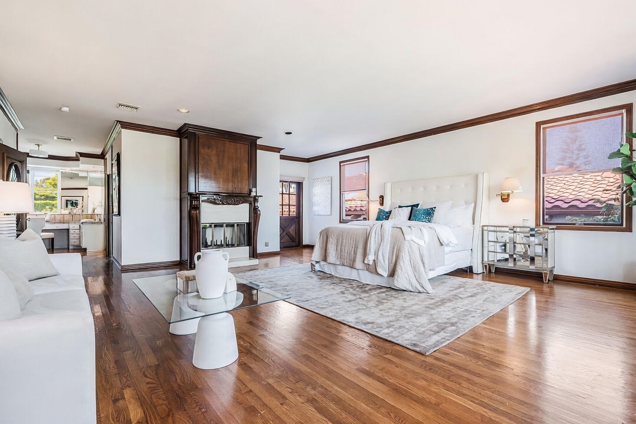 This spacious primary bedroom features warm wood flooring and a neutral color palette, centered around a large bed with a tufted headboard. A prominent wood-paneled fireplace surround serves as a focal point, while a glass-topped coffee table sits on a textured area rug in the foreground. The room is brightened by large windows and flows into an adjacent bathroom area, creating an open and airy atmosphere.