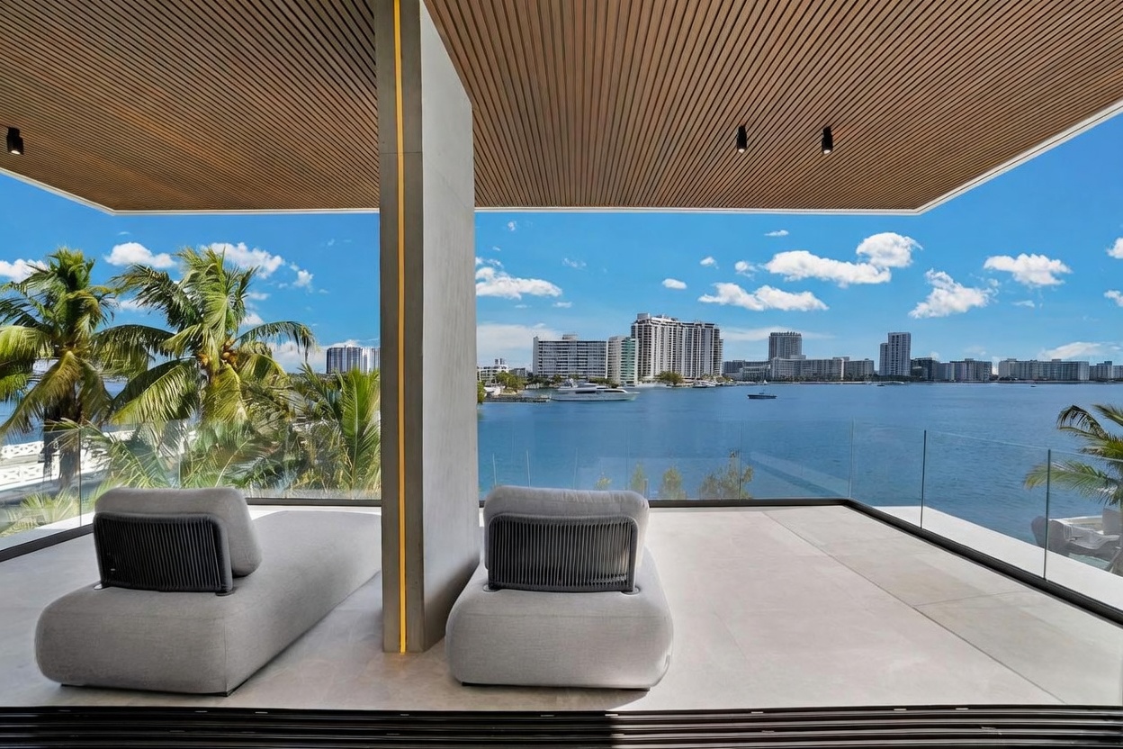 This luxurious balcony offers a panoramic view of the bay and city skyline, featuring two modern, low-profile lounge chairs positioned on a sleek, light-toned tiled floor. A central concrete pillar with integrated vertical lighting divides the space, while a frameless glass railing ensures an unobstructed view of the water and distant buildings. The area is sheltered by a slatted wood-look ceiling, creating a sophisticated and serene outdoor retreat.