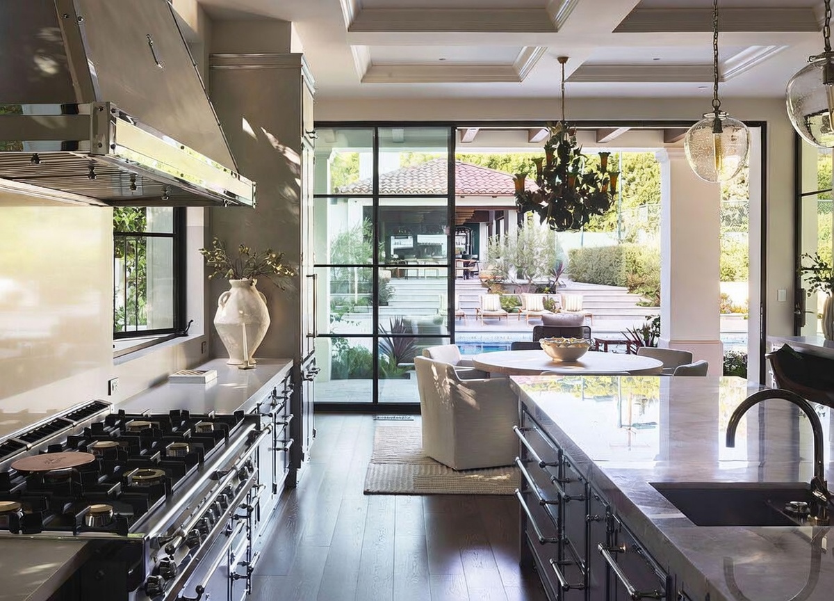 This sophisticated kitchen features a professional-grade range with a large metal hood, dark cabinetry, and a spacious stone-topped island. The space seamlessly transitions to an outdoor patio through expansive floor-to-ceiling glass doors, creating an airy, indoor-outdoor living experience. A dining area is positioned near the glass wall, offering a clear view of the pool and landscaped backyard, while elegant pendant lighting adds a refined touch to the open-concept design.