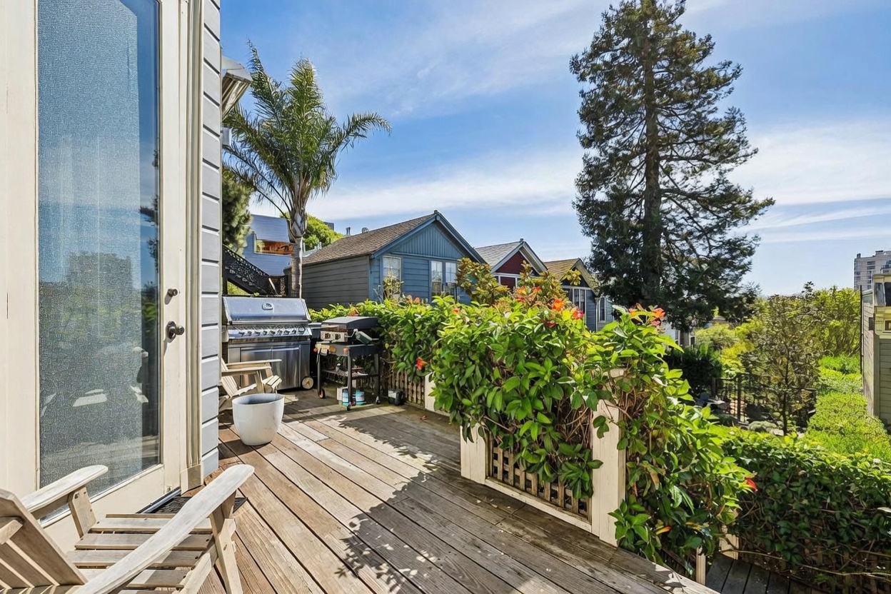 This elevated wooden deck offers a private outdoor living space with a grill setup and a view of the surrounding neighborhood. Lush greenery and flowering plants line the railing, creating a vibrant and inviting atmosphere. The perspective captures the deck from a doorway, highlighting the seamless transition between indoor and outdoor living areas.