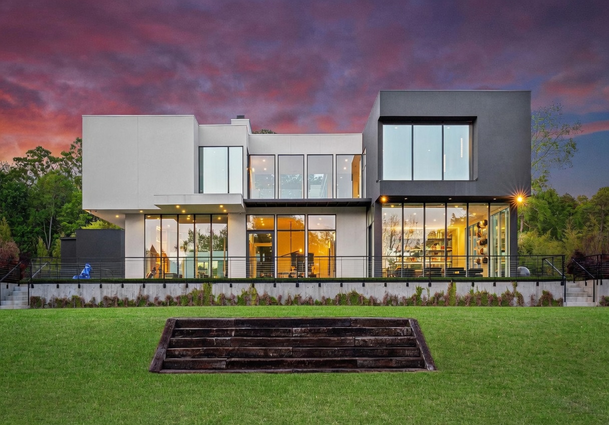 This striking modern residence features a bold, geometric design with a contrasting palette of light gray and dark charcoal exterior panels. The rear facade is defined by extensive glass walls that provide a seamless connection between the interior living spaces and the expansive backyard. A raised terrace with a sleek metal railing overlooks a manicured lawn, which is accessed by a set of wide, rustic timber steps.
