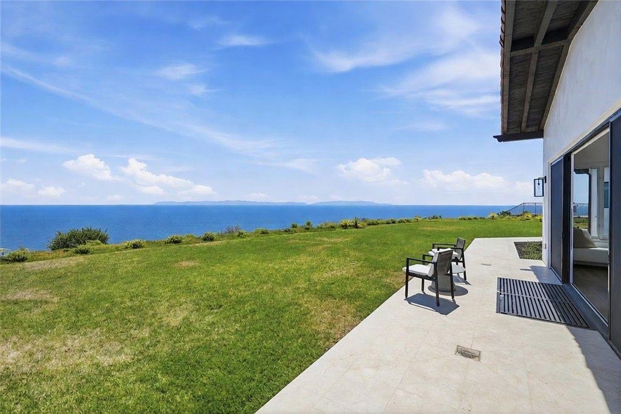 This image captures a spacious, light-colored stone patio that transitions seamlessly into a vast, manicured lawn overlooking the ocean. Two comfortable outdoor chairs are positioned on the patio, providing a perfect vantage point for the expansive coastal view and distant islands. The scene is framed by a clear blue sky and the clean lines of the home's exterior, creating a serene and luxurious atmosphere.