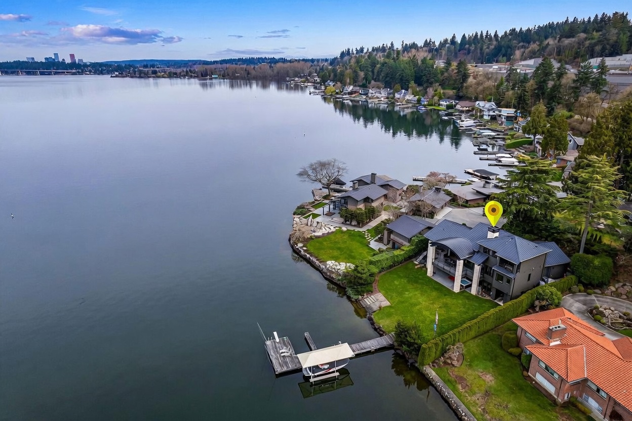 This high-angle aerial view captures a luxurious waterfront property situated on a point, featuring a modern home with a dark metal roof and a private dock extending into the calm lake. The expansive lawn slopes gently toward the water, framed by mature trees and neighboring residences along the shoreline. In the distance, a city skyline is visible across the water, providing a striking contrast between the serene natural setting and urban development.