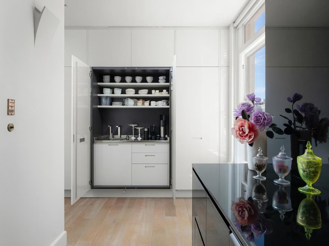 This interior view showcases a sleek, modern kitchenette concealed within a wall of white cabinetry. The open cabinet reveals a stainless steel countertop, a small sink, and organized shelving filled with dishware and coffee accessories. To the right, a black glossy surface holds decorative glass jars and a vase of flowers, while light wood flooring grounds the minimalist space.