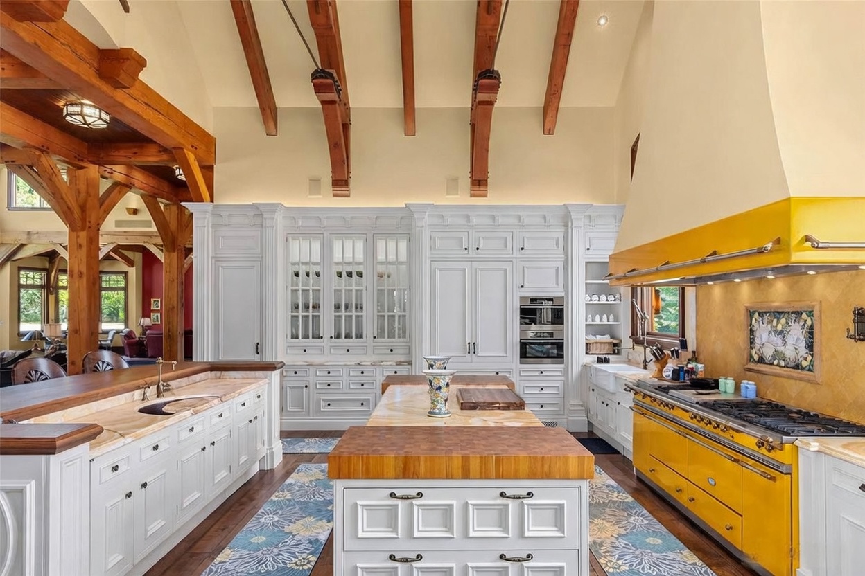 This expansive, high-ceilinged kitchen features a striking contrast between white cabinetry and a vibrant yellow range and hood. A large central island with a thick wood-block top serves as a focal point, complemented by stone-topped counters and exposed timber beams overhead. The perspective is a wide, symmetrical shot looking directly into the heart of the kitchen, showcasing the blend of rustic architectural elements and colorful, high-end appliances.