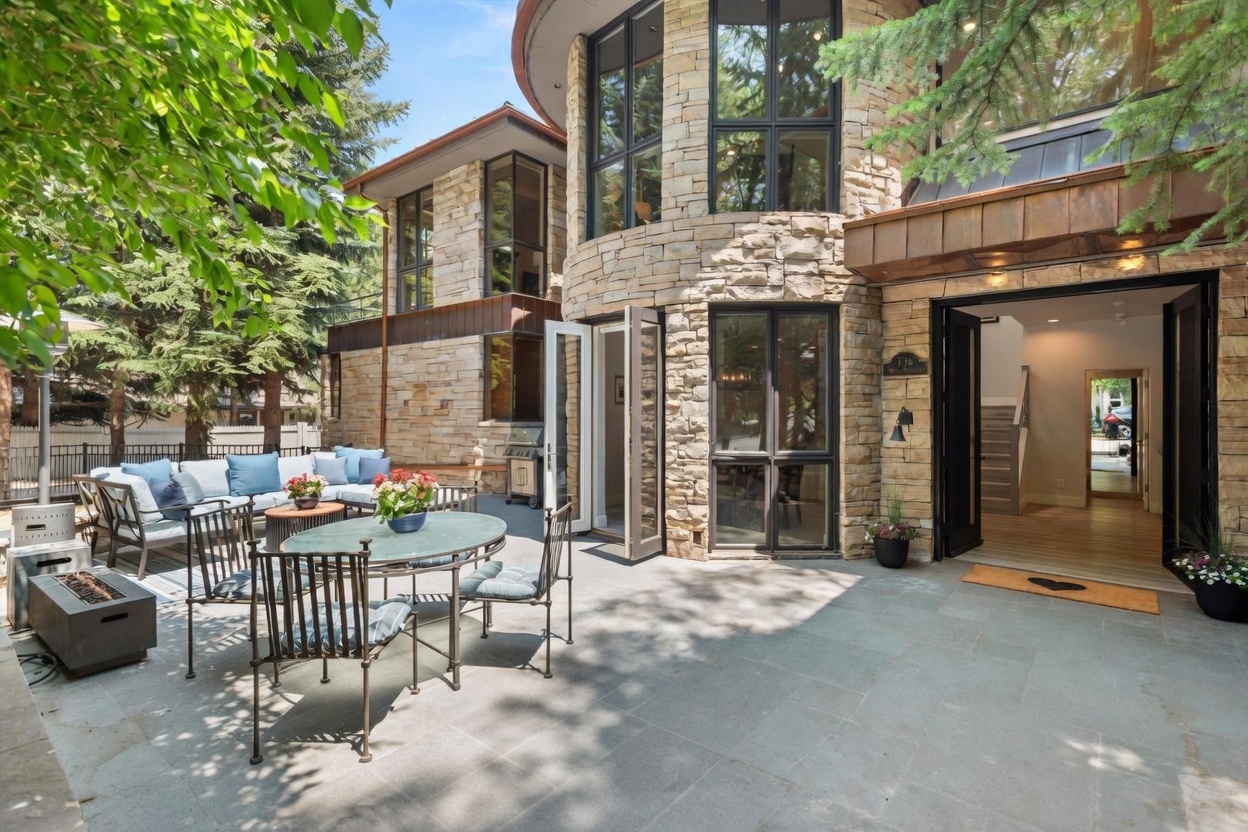 This inviting stone-paved patio features a comfortable outdoor seating area with plush blue cushions and a glass-topped dining set, perfect for al fresco entertaining. The home's exterior showcases a striking curved stone facade with large windows and a copper-accented roofline, blending rustic textures with modern architectural design. Lush greenery and vibrant potted flowers frame the space, creating a serene and private atmosphere.