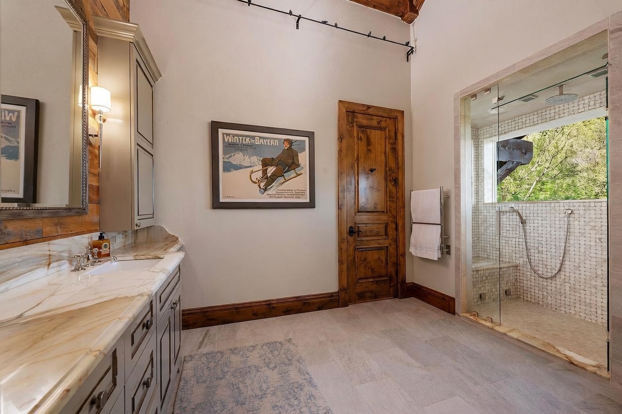 This primary bathroom features a vanity with a stone countertop and a large mirror, complemented by a walk-in glass-enclosed shower with a window view. The room is decorated with a vintage-style ski poster, warm wood-toned doors, and neutral tile flooring, creating a rustic yet refined atmosphere. The perspective is a wide-angle shot capturing the transition from the vanity area to the shower space.