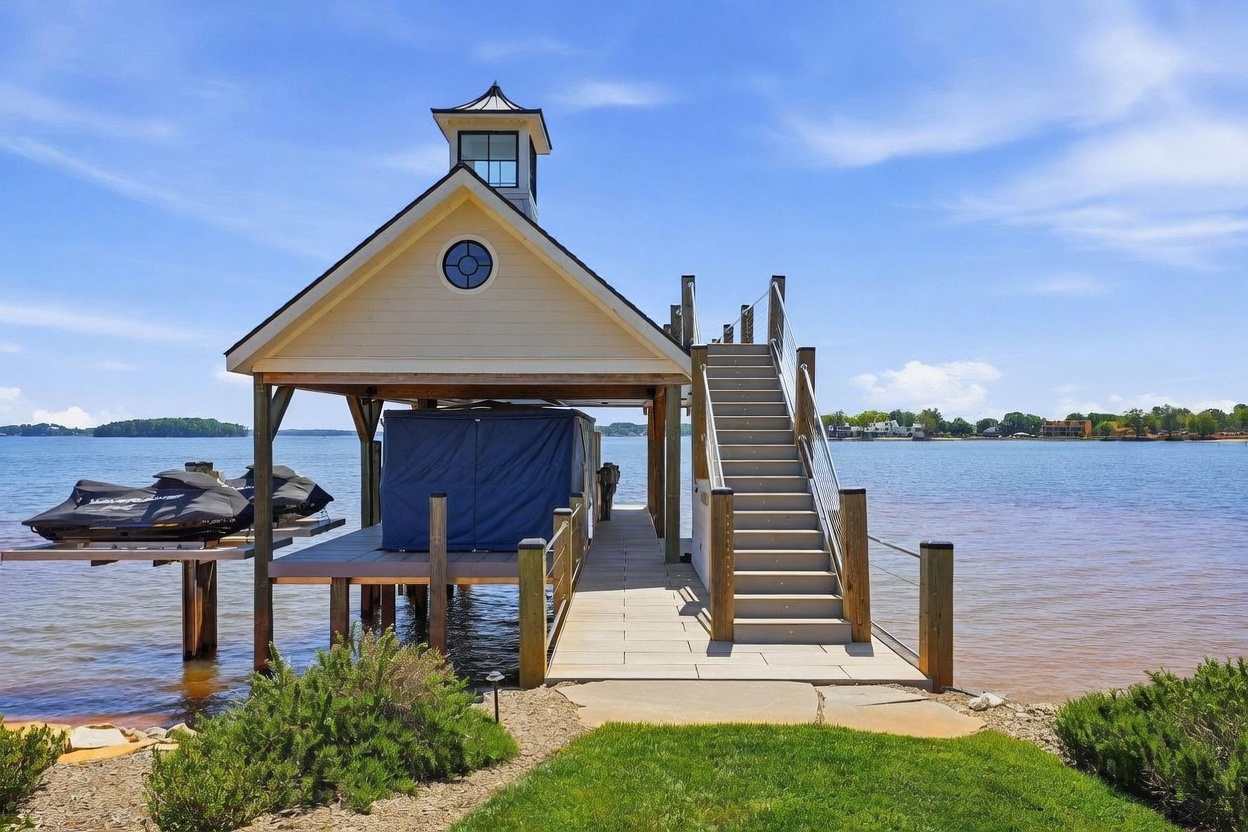 This image captures a private boat dock and covered slip extending into a calm, expansive lake. The structure features a light-colored exterior with a cupola, a wooden walkway, and a staircase leading to an upper deck area, with two personal watercraft docked underneath. The scene is set under a bright blue sky, emphasizing the recreational lifestyle and waterfront access provided by the property.