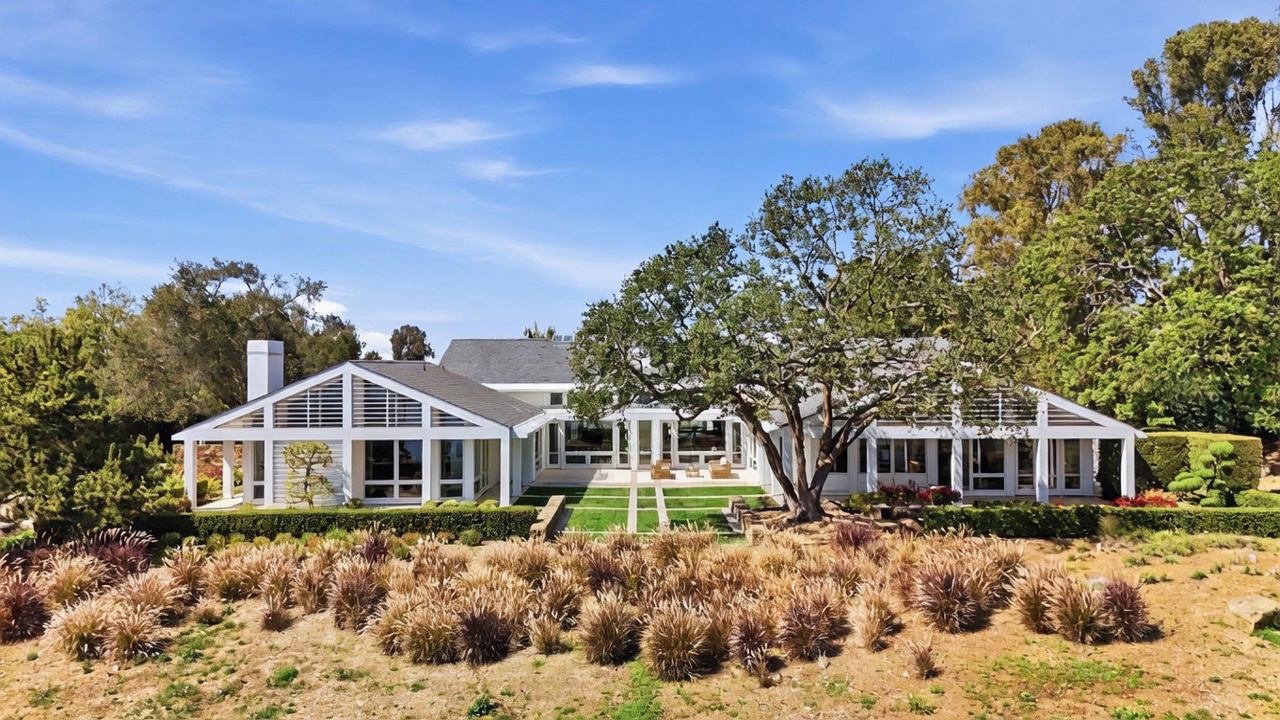 This wide-angle exterior shot captures the rear elevation of a contemporary residence, characterized by its expansive glass walls and clean, geometric lines. A large, mature oak tree serves as a focal point in the center of the manicured lawn, which features a grid of stone pathways. The property is surrounded by lush greenery and native landscaping, creating a serene and private atmosphere.