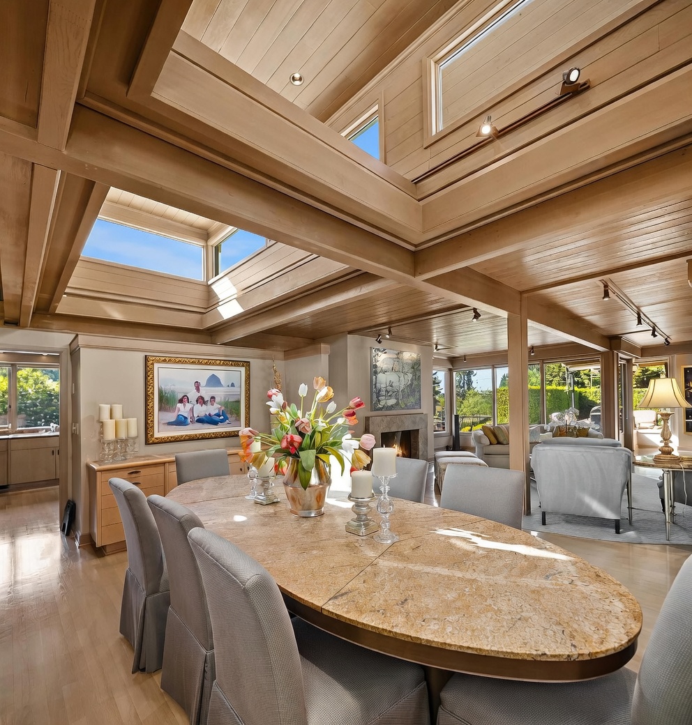 This elegant dining area features a large oval table with a stone top, surrounded by upholstered chairs, positioned beneath a dramatic vaulted ceiling with multiple skylights. The open-concept space flows into a living area with a stone-clad fireplace and large windows that provide abundant natural light. The room is finished with light wood flooring and warm wood ceiling beams, creating a sophisticated and airy atmosphere.