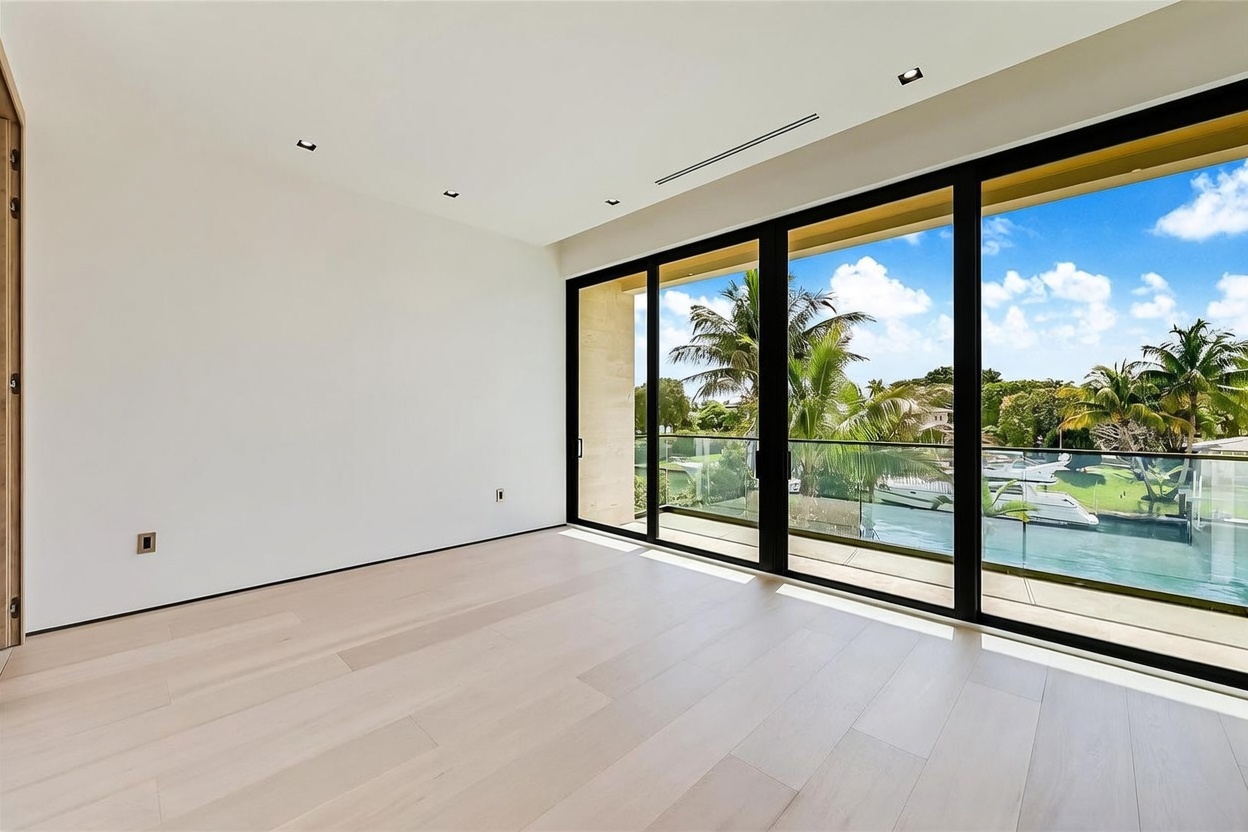 This bright, minimalist room features light wood flooring and crisp white walls, creating an airy and open atmosphere. A wall of floor-to-ceiling glass sliding doors provides seamless access to a balcony and offers expansive views of the water and surrounding palm trees. The space is clean and modern, emphasizing natural light and a connection to the outdoor environment.