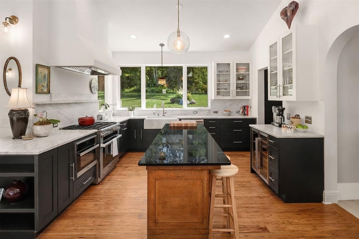 This spacious kitchen features a striking contrast between dark lower cabinetry and white upper cabinets, complemented by a large central island with a dark stone countertop and a warm wood base. A wide window above the farmhouse sink provides a clear view of the lush green backyard, while elegant pendant lighting hangs above the island. The room is finished with light wood flooring and stone countertops, creating a sophisticated and inviting culinary space.