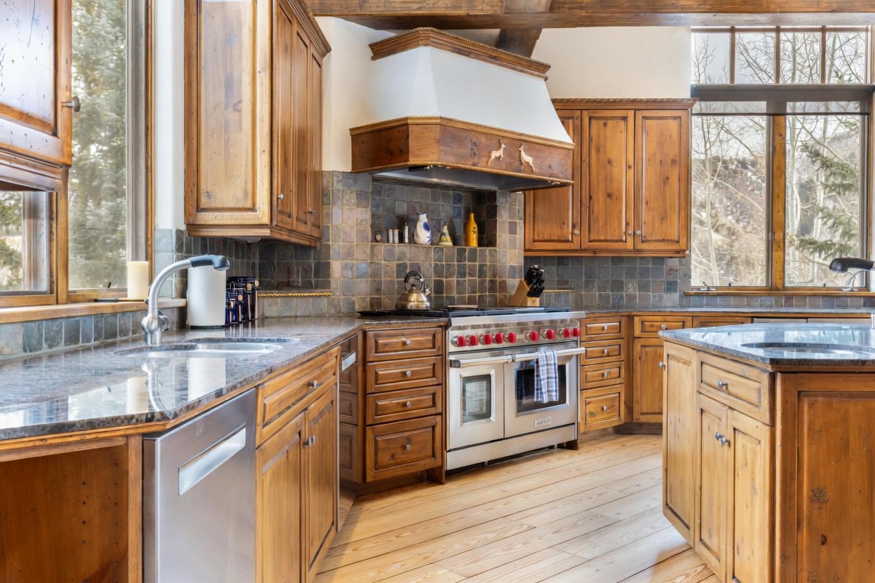 This rustic-style kitchen features warm-toned wood cabinetry, dark stone countertops, and a professional-grade stainless steel range with a prominent hood. The space is accented by a tiled backsplash and large windows that provide ample natural light and views of the outdoors. The perspective captures the functional layout, highlighting the island, the integrated appliances, and the cohesive, mountain-inspired aesthetic.