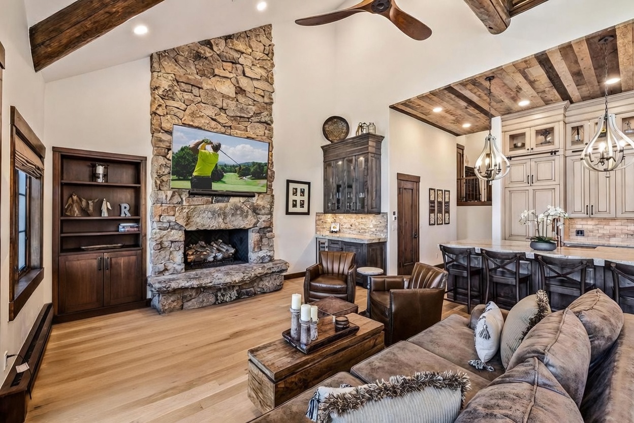 This expansive living room features a rustic aesthetic with a prominent stone-clad chimney breast housing a television above the fireplace. The open-concept space includes a cozy seating area with leather armchairs and a plush sofa, transitioning into a kitchen area with a breakfast bar and wooden ceiling beams. The warm lighting and natural textures create an inviting, lodge-inspired atmosphere.