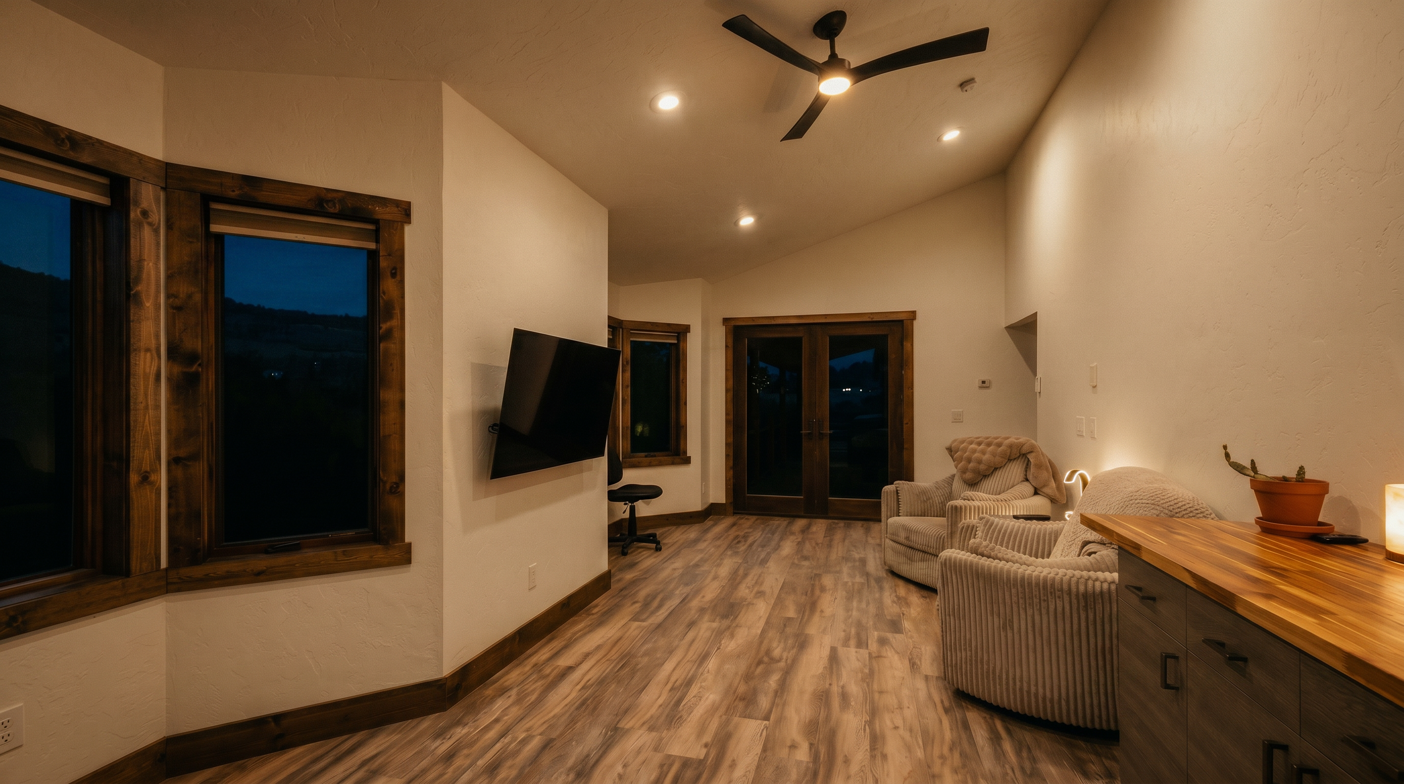 This bright living area features light wood-look flooring and white walls, creating an airy and open atmosphere. The space is furnished with two comfortable armchairs and includes a wall-mounted television, while large windows and glass doors provide expansive views of the surrounding landscape. A ceiling fan centers the room, and a partial view of a cabinet with a wood-toned countertop adds a functional touch to the layout.